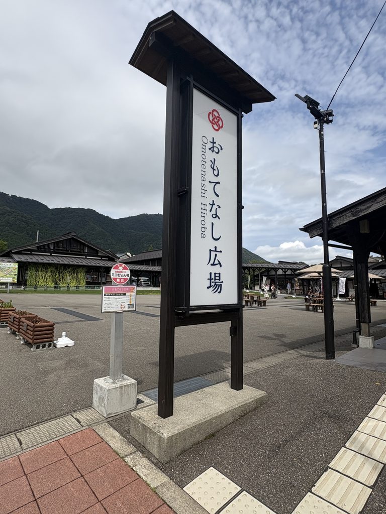 おもてなし広場の看板。テーブルとパラソルが並ぶ広場